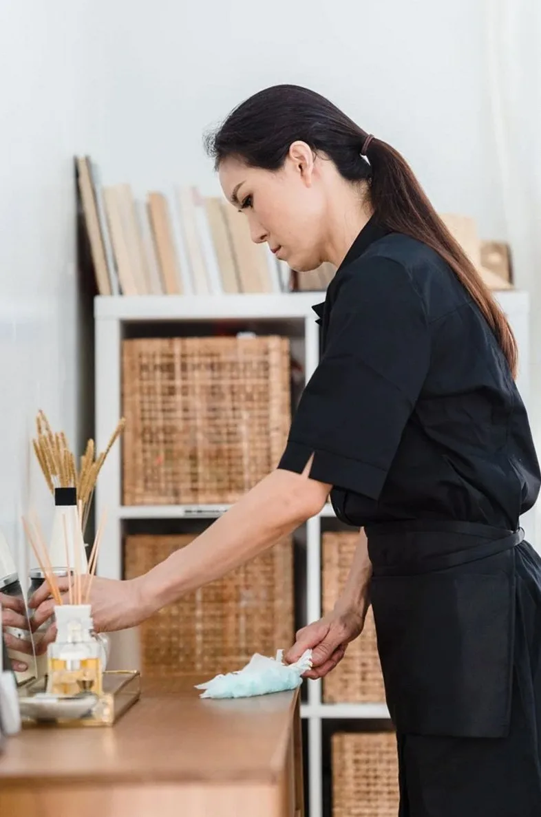 黒い制服を着た女性が室内で木製のカウンターを拭いており、背景に棚が並んでいる。