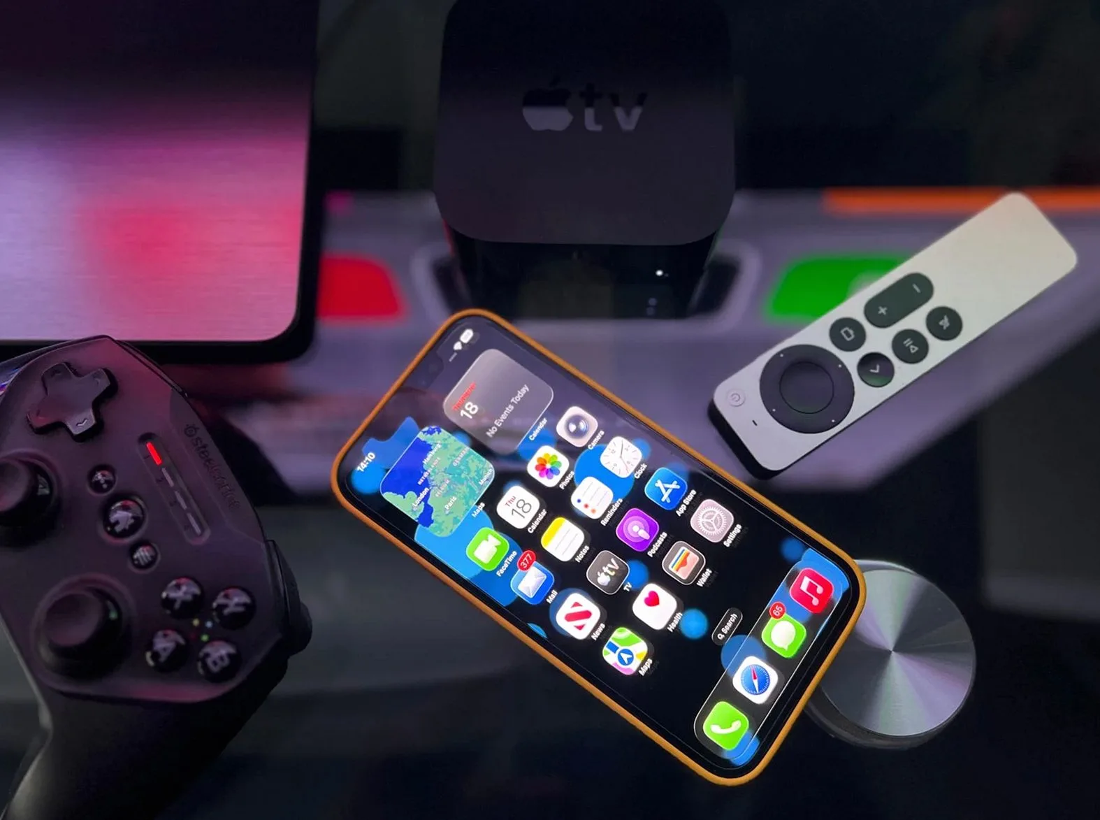 ゲームコントローラー、iPhone、Apple TV本体とリモコンが並ぶ暗めのデスク